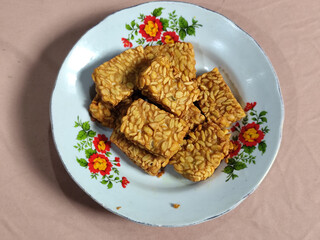 A pile of crispy fried tempeh that looks delicious and tasty, in a glass plate with a floral motif.
