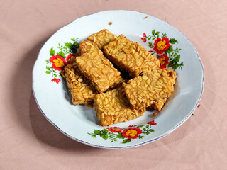 A pile of crispy fried tempeh that looks delicious and tasty, in a glass plate with a floral motif.