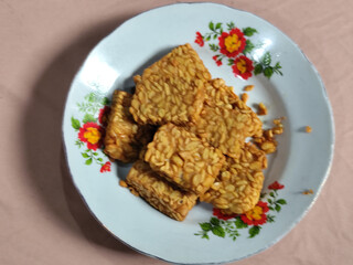 A pile of crispy fried tempeh that looks delicious and tasty, in a glass plate with a floral motif.
