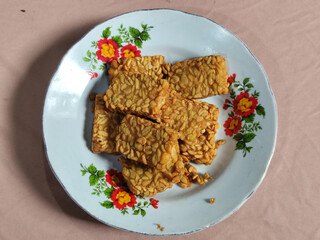 A pile of crispy fried tempeh that looks delicious and tasty, in a glass plate with a floral motif.
