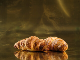 Two croissants, fresh and golden, sit invitingly on a shiny surface. Morning light catches their delicate curves, tempting with the promise of a delicious treat