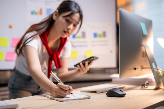 Asian businesswoman taking notes and using smartphone in the office - Powered by Adobe