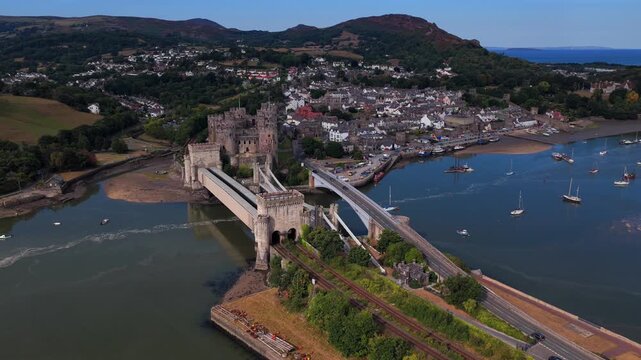 Aerial video of Conwy Castle, railway line, and road bridge crossing the estuary with the town and mountains in the background on a summer day.

