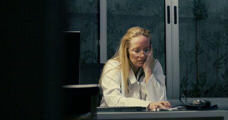 Female doctor sitting at desk with head down holding face in hands under office light in late hours...