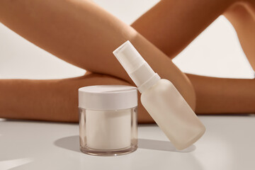 White cream jar and spray bottle on table near crossed legs of woman closeup. Concept of skincare set, beauty lifestyle, wellness routine, cosmetic advertising, and luxury feminine care.