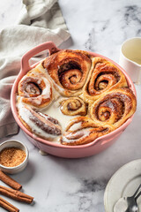 Freshly baked glazed cinnabon buns in a ceramic baking dish on white marble background. Making cinnamon rolls process