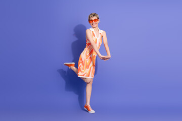 Obraz premium Stylish young woman in vibrant patterned dress and sunglasses posing against a solid purple background in a cheerful mood