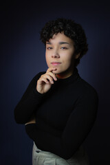 Beautiful young woman with short curly hair,  wearing black turtleneck blouse, looking at camera, hand on chin. Studio portrait, copy space on dark background. Ideal for Gen Z attitude, contemplation.