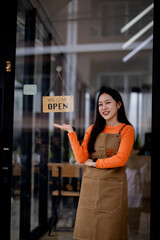 Cheerful male café owner in apron standing at store entrance, smiling proudly with “open” sign, symbolizing success and welcoming customers.
