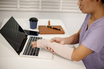 A person in a purple shirt sits at a desk, holding a blue smartphone in their left hand, while their right arm, wrapped in a beige cast, rests beside an open laptop