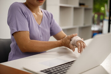 A person in a purple shirt sits at a desk, holding a blue smartphone in their left hand, while their right arm, wrapped in a beige cast, rests beside an open laptop