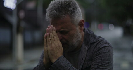 Middle aged man with eyes shut and hands clasped in desperate prayer or emotional plea, standing in rain