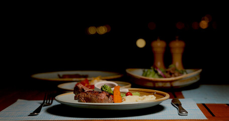 Evening dining table with steak, mashed potatoes, vegetables. Cozy restaurant ambiance, warm lighting, delicious meal prepared for elegant dinner.