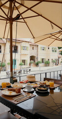 Charming outdoor cafe breakfast setup under umbrella in picturesque European village. Plates filled with delicious food, vibrant atmosphere, inviting dining experience.