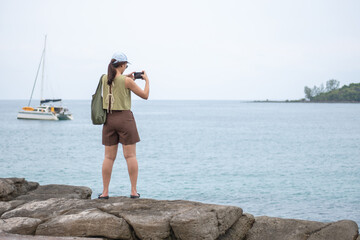 Asian woman taking photo of sailboat on ocean from rocky shore during daytime