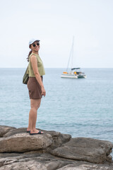 Young Asian woman standing on rocks by the sea with sailboat in background