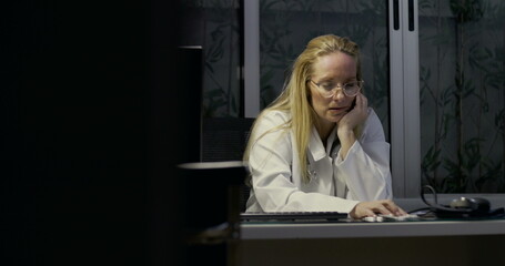 Female doctor holding face in hands at desk appearing mentally and physically exhausted during late hours in medical clinic