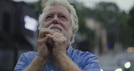 Elderly man holding clasped hands near face, eyes glistening in rain, filled with emotion, hope, or silent prayer in moment of spiritual connection