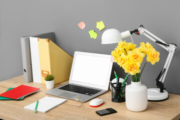 Workplace with bouquet of narcissus flowers, lamp and laptop near black wall in stylish office