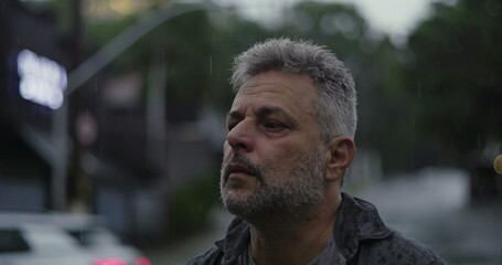 Middle aged man with serious downward gaze, drenched in rain, expressing sorrowful reflection and emotional weight