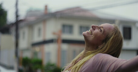 Blonde woman smiling joyfully in residential street under rain, looking up with emotional release...