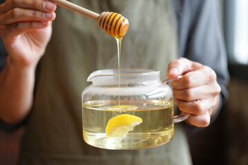 Close-up of hands holding a glass teapot with steaming hot lemon tea while golden honey drips from a wooden dipper, creating a cozy and natural wellness drink moment