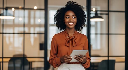 A smiling african american woman with curly hair holding a tablet and standing in a modern office with glass partitions