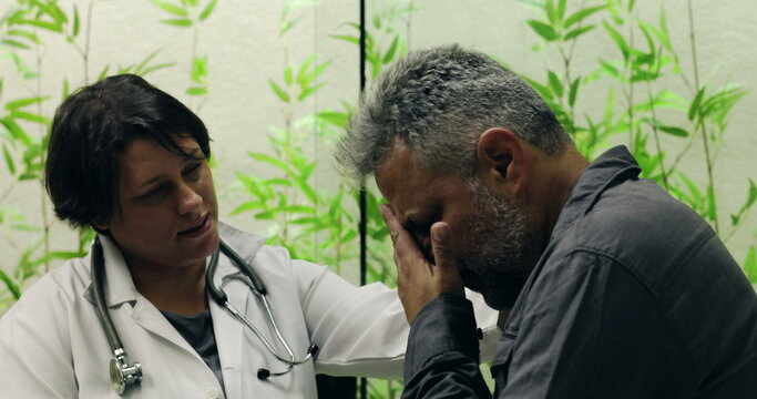Female doctor speaking gently to distressed middle-aged male patient during emotional conversation in clinic, offering support with hand on shoulder