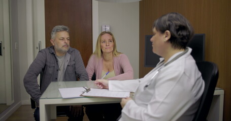 Fototapeta premium Doctor consulting middle-aged couple during appointment at clinical office, clipboard and paperwork visible on desk