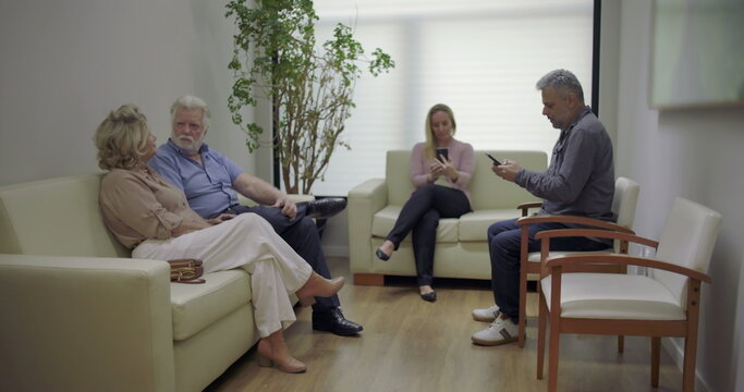 Group of patients in clinic waiting area, including elderly couple talking and others using phones, seated on couches and chairs in calm setting - Powered by Adobe