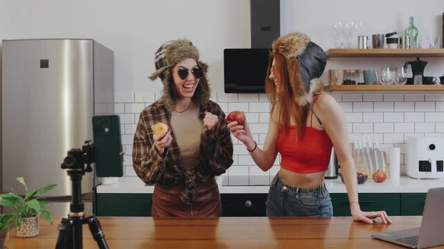Two bloggers in fuzzy hats film a hilarious kitchen video, racing to eat apples as fast as possible while laughing through bites and reacting with playful exaggeration