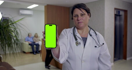 Doctor holding smartphone with green screen facing camera in hospital hallway while nurse and patients are visible near reception and waiting area