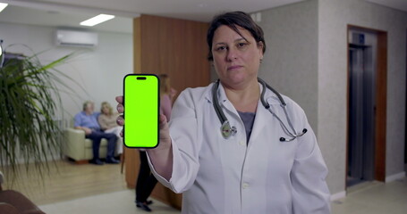 Doctor holding smartphone with green screen facing camera in hospital hallway while nurse and patients are visible near reception and waiting area