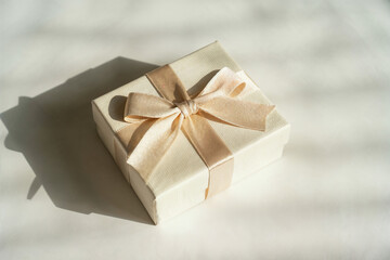 Obraz premium Gift Box with Ribbon on Fabric. A small beige gift box with a satin ribbon placed on soft fabric with natural shadows. Suitable for celebrations, weddings, and holiday concepts.
