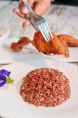 Adult hand holding a crispy fried chicken wing with a fork and spoon, prepared for serving alongside a mound of wholesome brown rice, a healthy Thai meal.