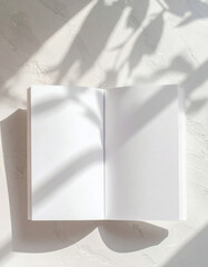An open blank notebook with white pages, casting shadows from a plant, on a textured surface.