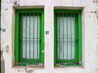 Two vibrant green windows with security bars are set into a heavily weathered and cracked white wall. A green graffiti symbol on the central pillar enhances the rustic, urban decay