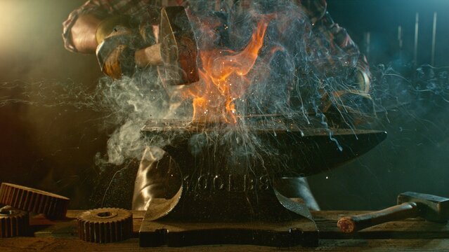 The blacksmith manually forging the molten metal on the anvil in smithy with spark fireworks