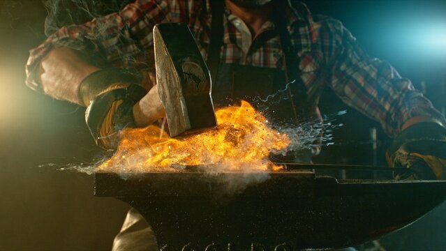 The blacksmith manually forging the molten metal on the anvil in smithy with spark fireworks