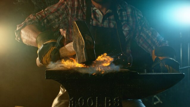 The blacksmith manually forging the molten metal on the anvil in smithy with spark fireworks