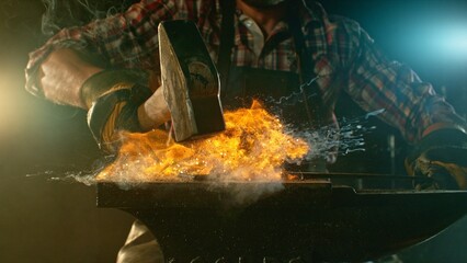 The blacksmith manually forging the molten metal on the anvil in smithy with spark fireworks