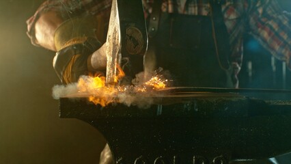 The blacksmith manually forging the molten metal on the anvil in smithy with spark fireworks