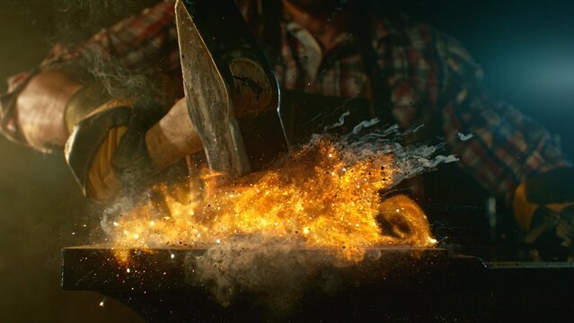 The blacksmith manually forging the molten metal on the anvil in smithy with spark fireworks