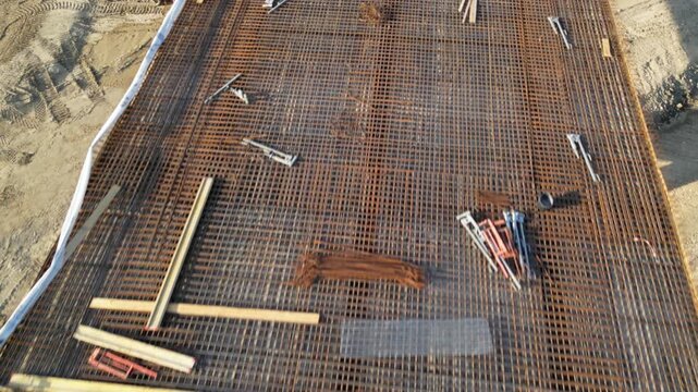 Top-down aerial view of a construction foundation covered with a dense steel reinforcement mesh, with timber formwork, tools and building materials prepared before concrete pouring.