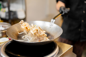 Chef stir-frying vegetables in a hot wok over a stove, wearing black gloves in a professional kitchen. Steam rises from the sizzling food, capturing dynamic cooking action.