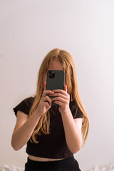 Young girl taking a selfie with a smartphone, standing in front of a light wall. Minimalist and candid lifestyle moment with soft natural lighting.