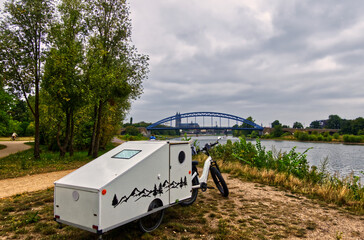White camper trailer hitched to electric bicycle, resting near elbe riverbank with historic sternbrücke bridge framing scenic magdeburg landscape