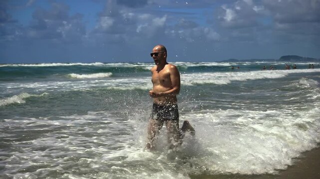 A hilarious middle-aged office escapee runs joyfully through ocean waves, his carefree sprint along a vast sandy beach under blue skies symbolizing the perfect break from routine.