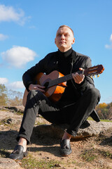 Beautiful blonde man playing acoustic guitar and singing relaxing enjoying outdoors on sunny autumn day. Artist learning and practicing playing musical instrument. Fall leaves autumnal weather 