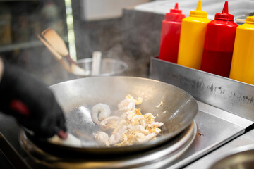 Chef stir-frying shrimp and scrambled eggs in a steaming wok at a commercial kitchen, wearing black gloves. Condiments and utensils visible in the background